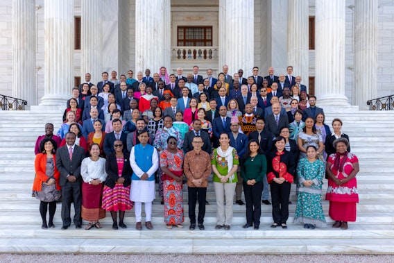 Group photograph of around 100 people in formal and cultural clothing standing on the steps of a white marble building, the Seat of the Universal House of Justice.