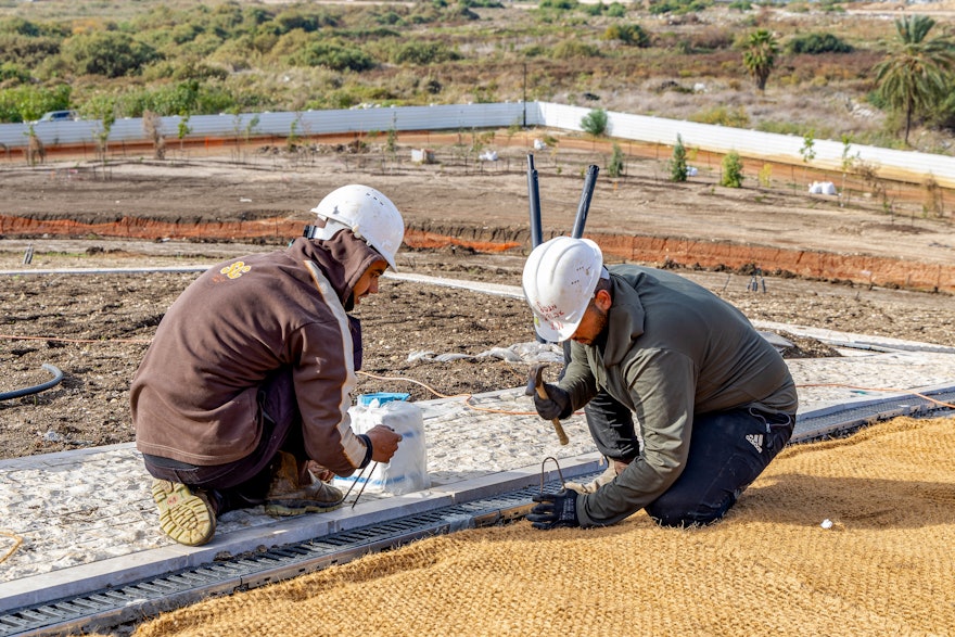 Workers on the west berm applying a waterproof layer made of coconut fiber for erosion control. Cobblestone paths have been completed, and installation of the irrigation system is next, enabling the landscaping to continue.