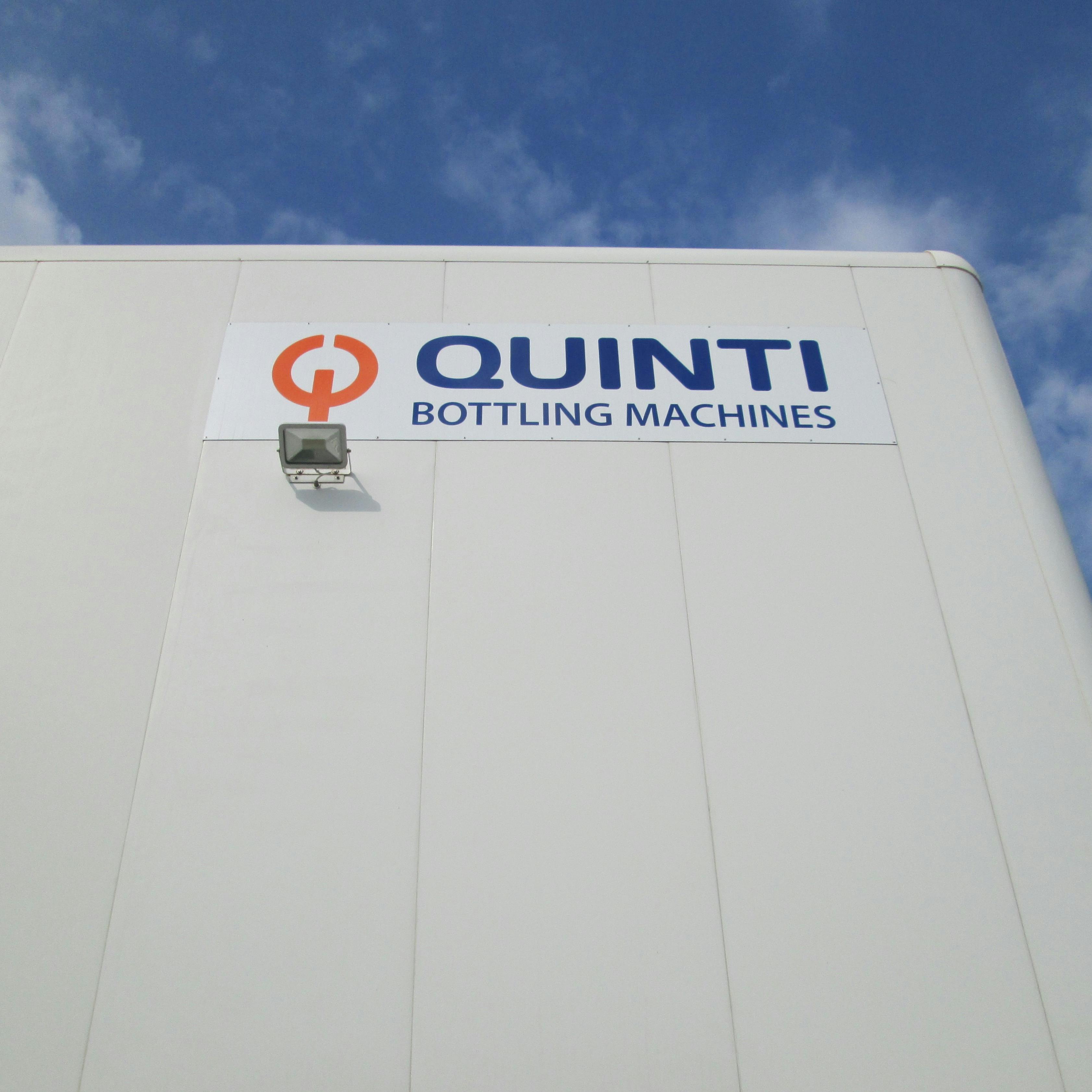 Insegna Quinti Bottling Machine con scritta di colore blu e logo di colore arancione, posata su capannone dell'azienda. Foto scattata dal basso verso l'alto. Sullo sfondo cielo colore azzurro con qualche nuvola bianca.