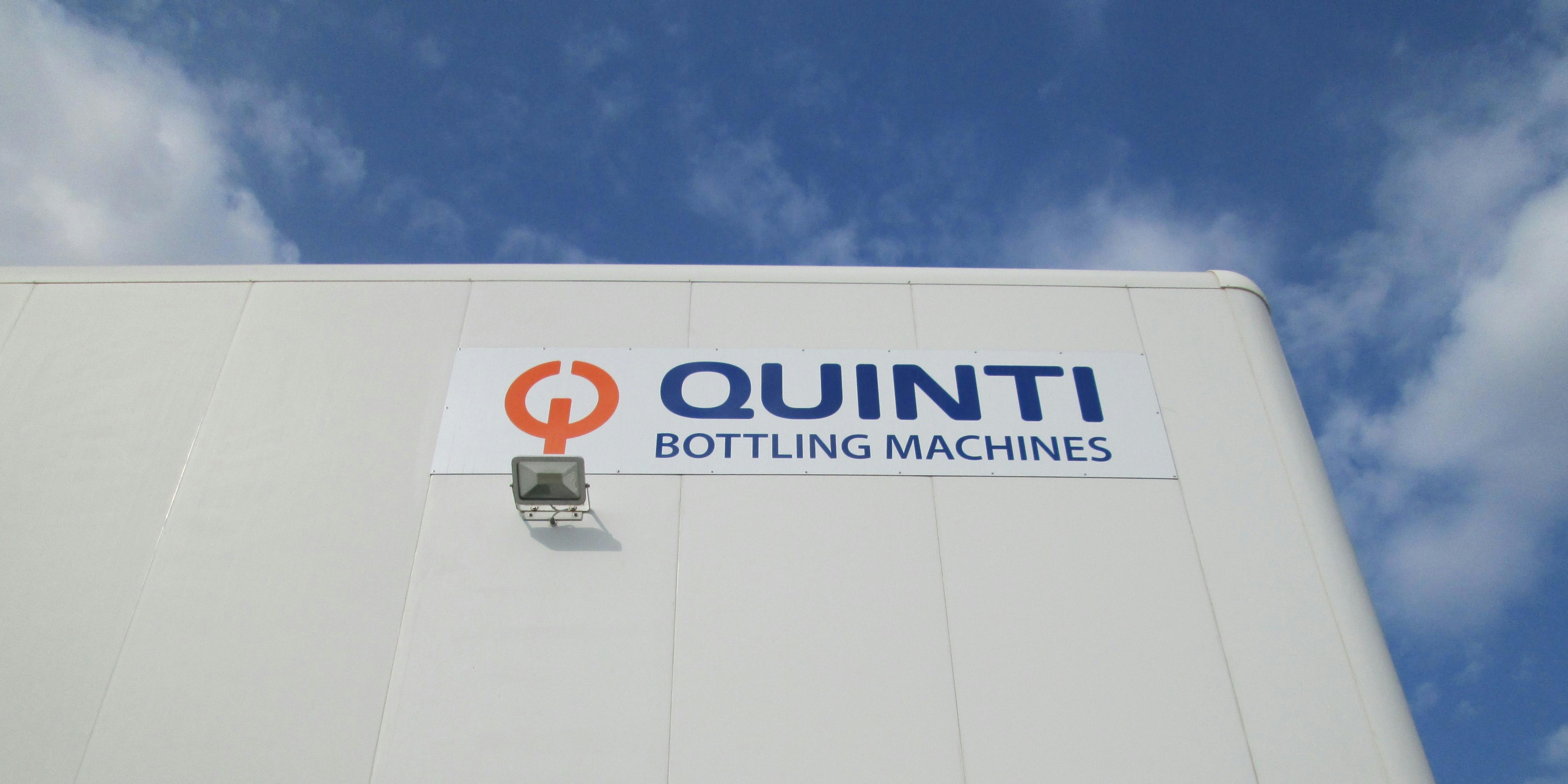 Quinti Bottling Machines sign whit blue writing and orange logo, placed on the company's headquarter. Photo taken from the bottom up. In the background blue sky with some white clouds.