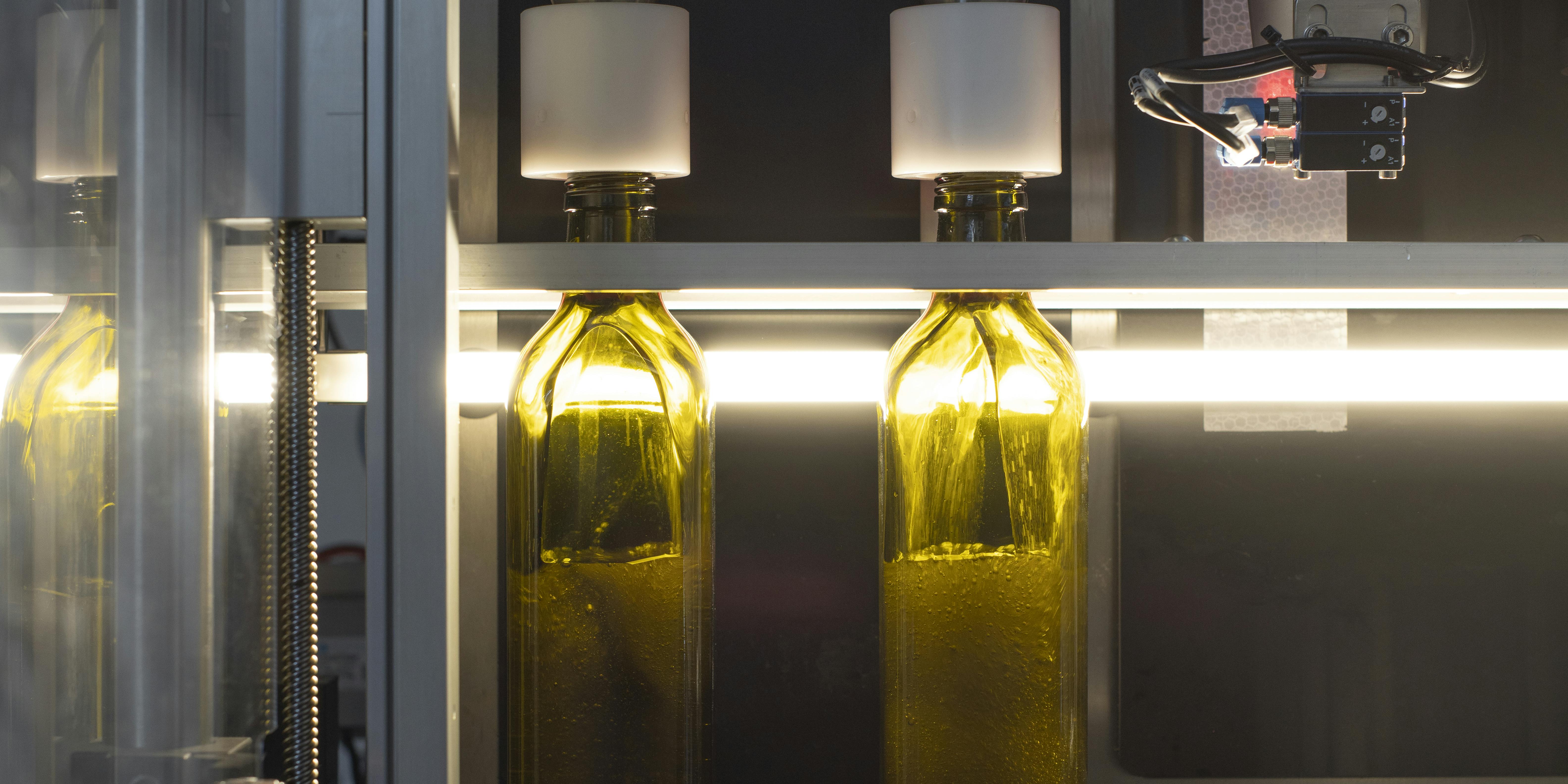 In questa foto è possibile vedere due bottiglie di olio quadre di colore verde, sostare nella stazione di rimpimento. L'olio entra nella bottiglia scivolando sulle sue pareti.