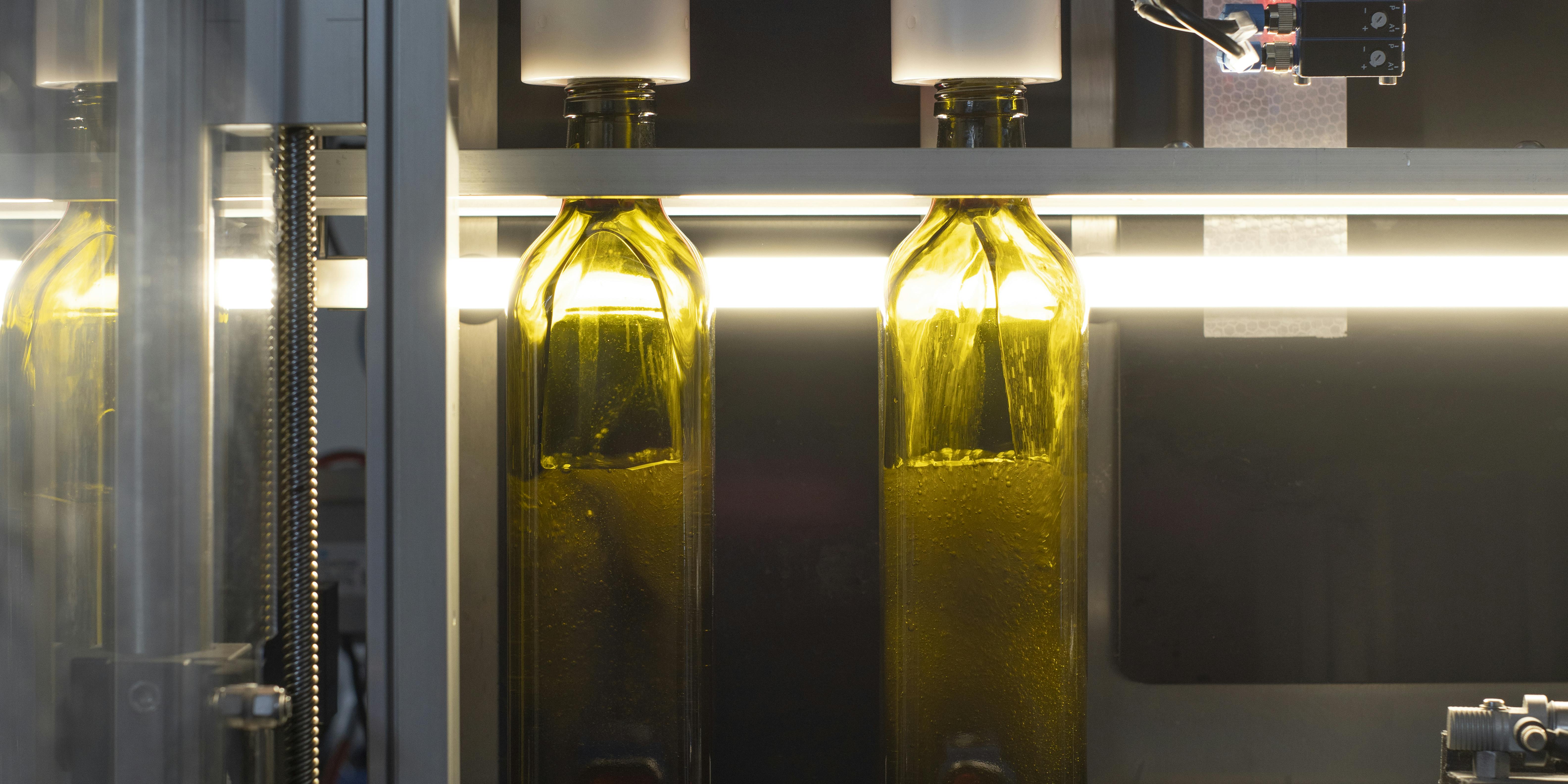 In questa foto è possibile vedere due bottiglie di olio quadre di colore verde, sostare nella stazione di rimpimento. L'olio entra nella bottiglia scivolando sulle sue pareti.