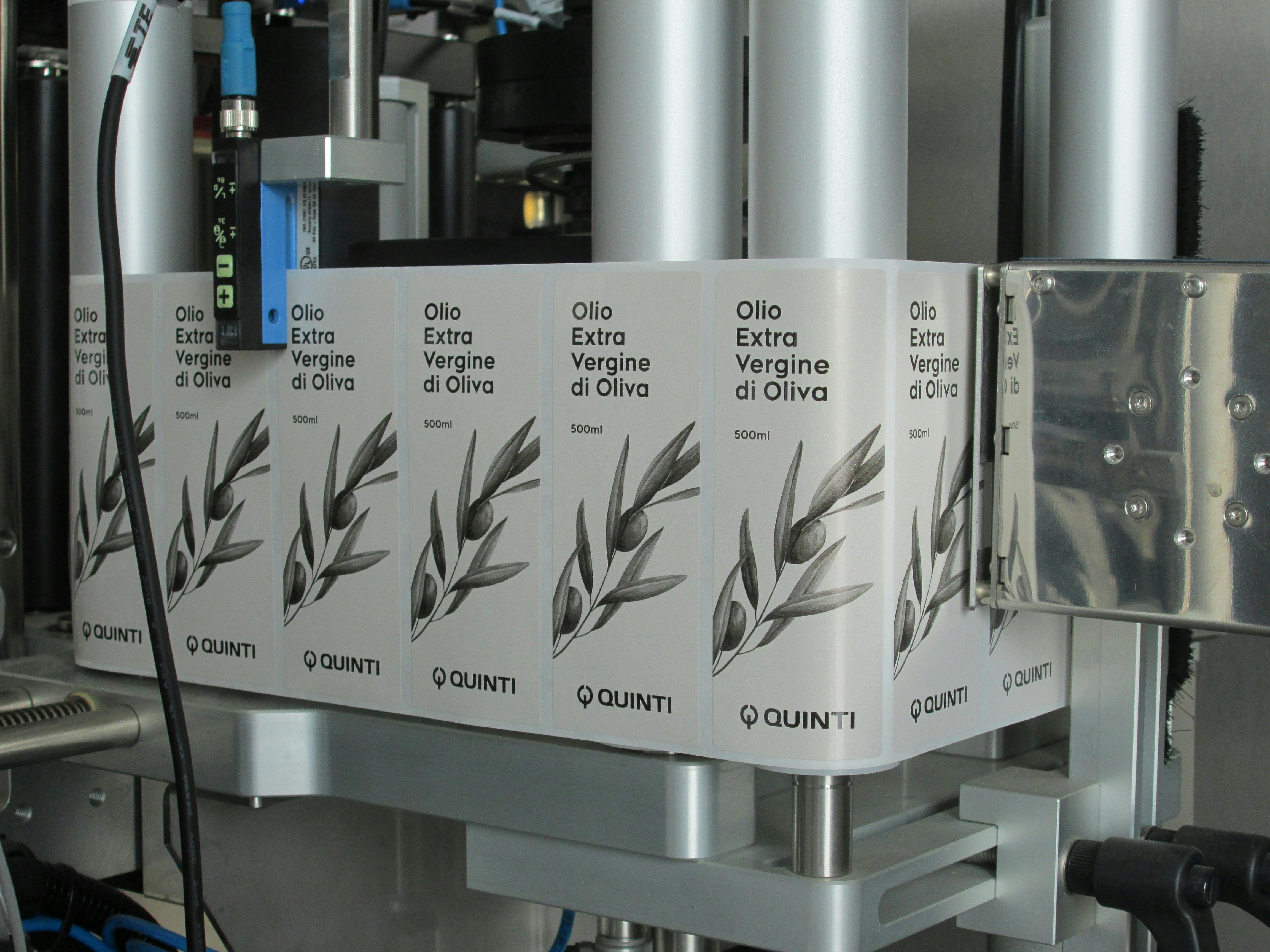 Dettaglio della macchina dove si vede etichettte rettangolari su bobina, indicate per bottiglia olio, raffiguranti ramo olivo e logo Quinti