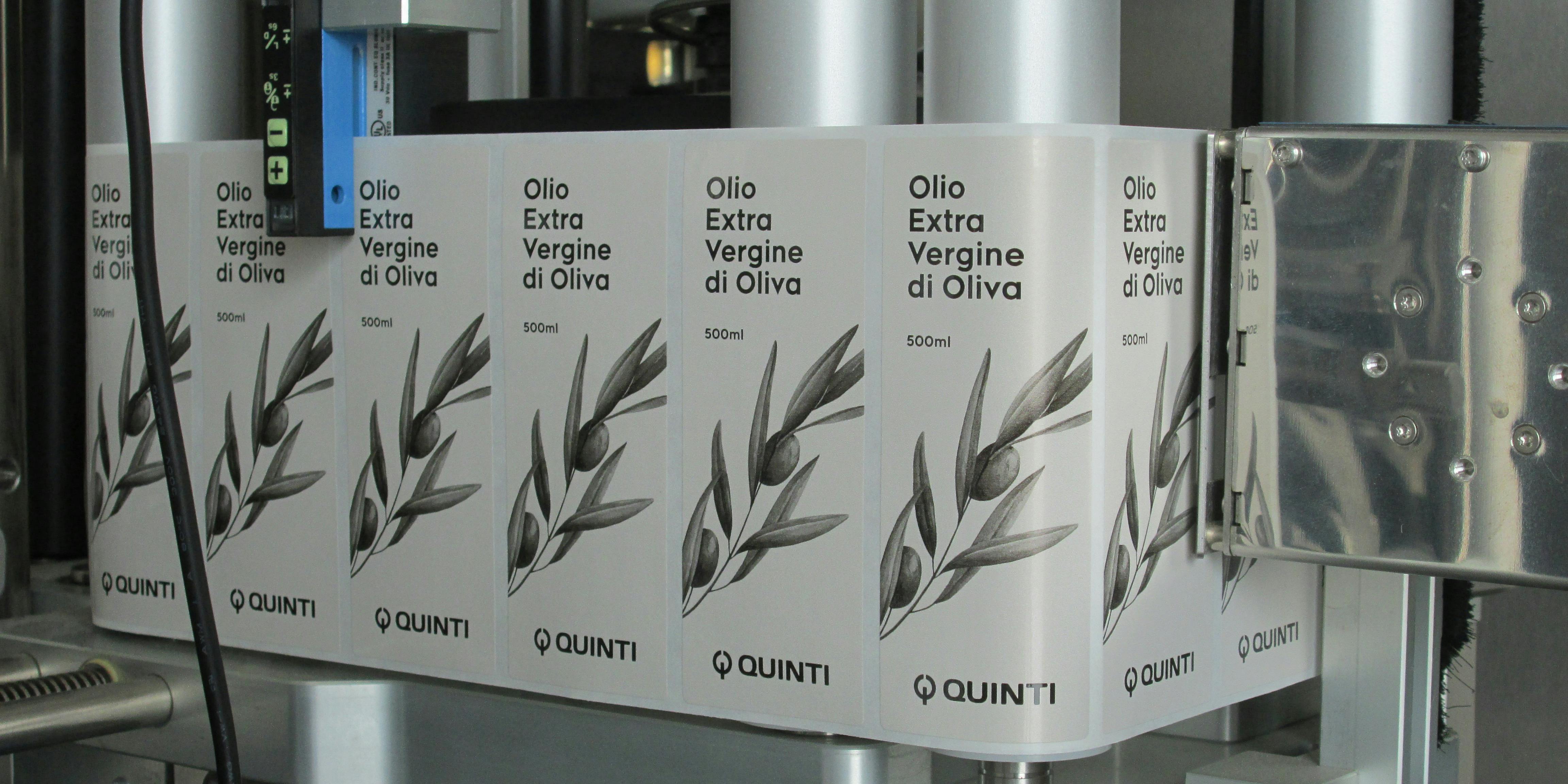Dettaglio della macchina dove si vede etichettte rettangolari su bobina, indicate per bottiglia olio, raffiguranti ramo olivo e logo Quinti