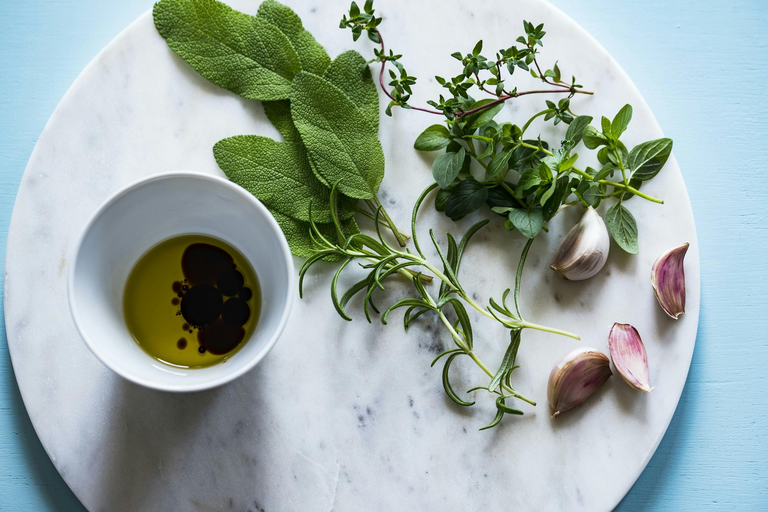 Un'immagine con un bicchiere di olio posato su marmo chiaro e al cui fiaco sono posizionate erbe aromatiche ed aglio