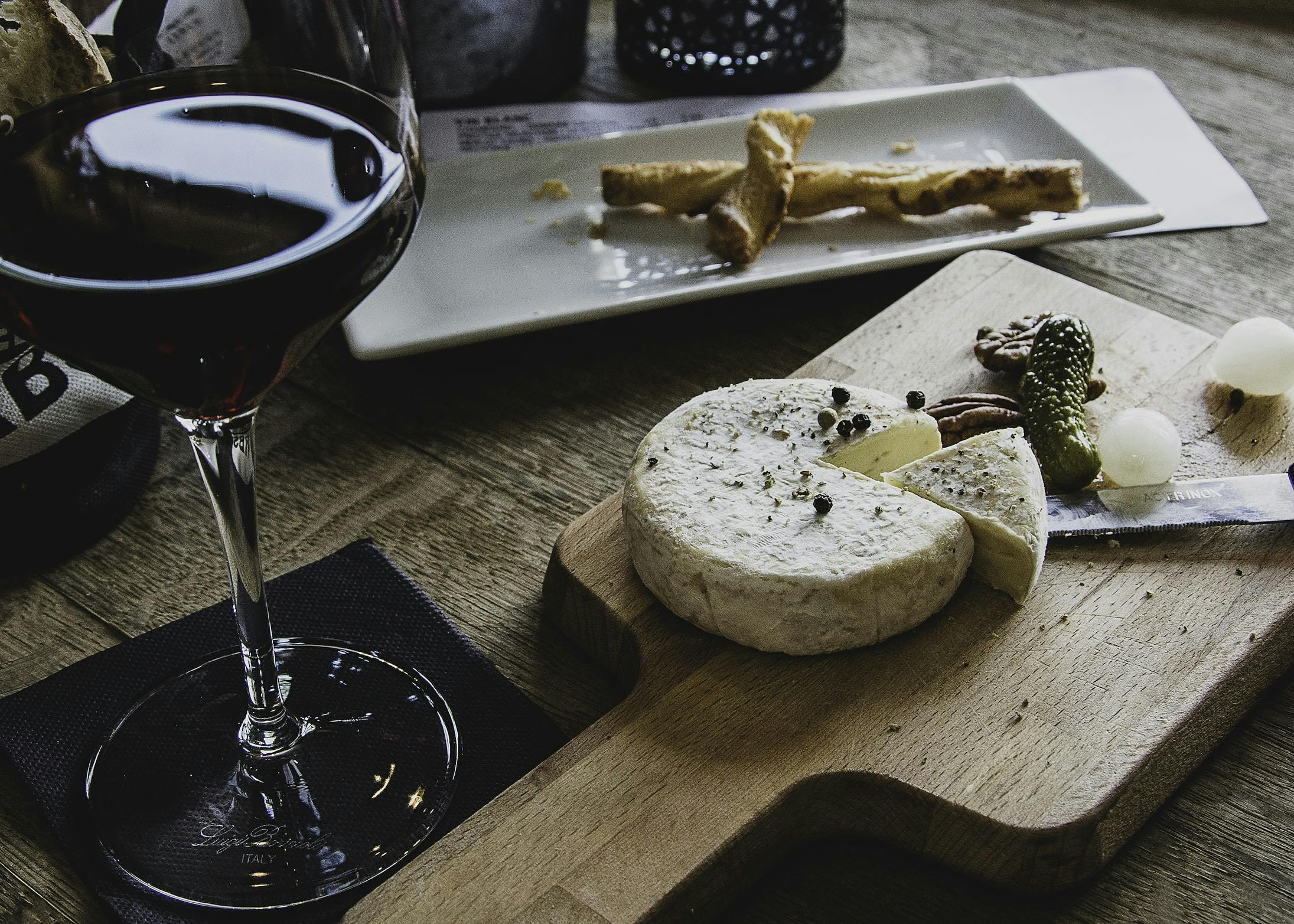 Un bicchiere di vino rosso, formaggio posato su di un tagliere di legno e nello sfondo un vassoio rettangolare di porcellana bianca su cui sono poste due sfoglie intrecciate di grissini
