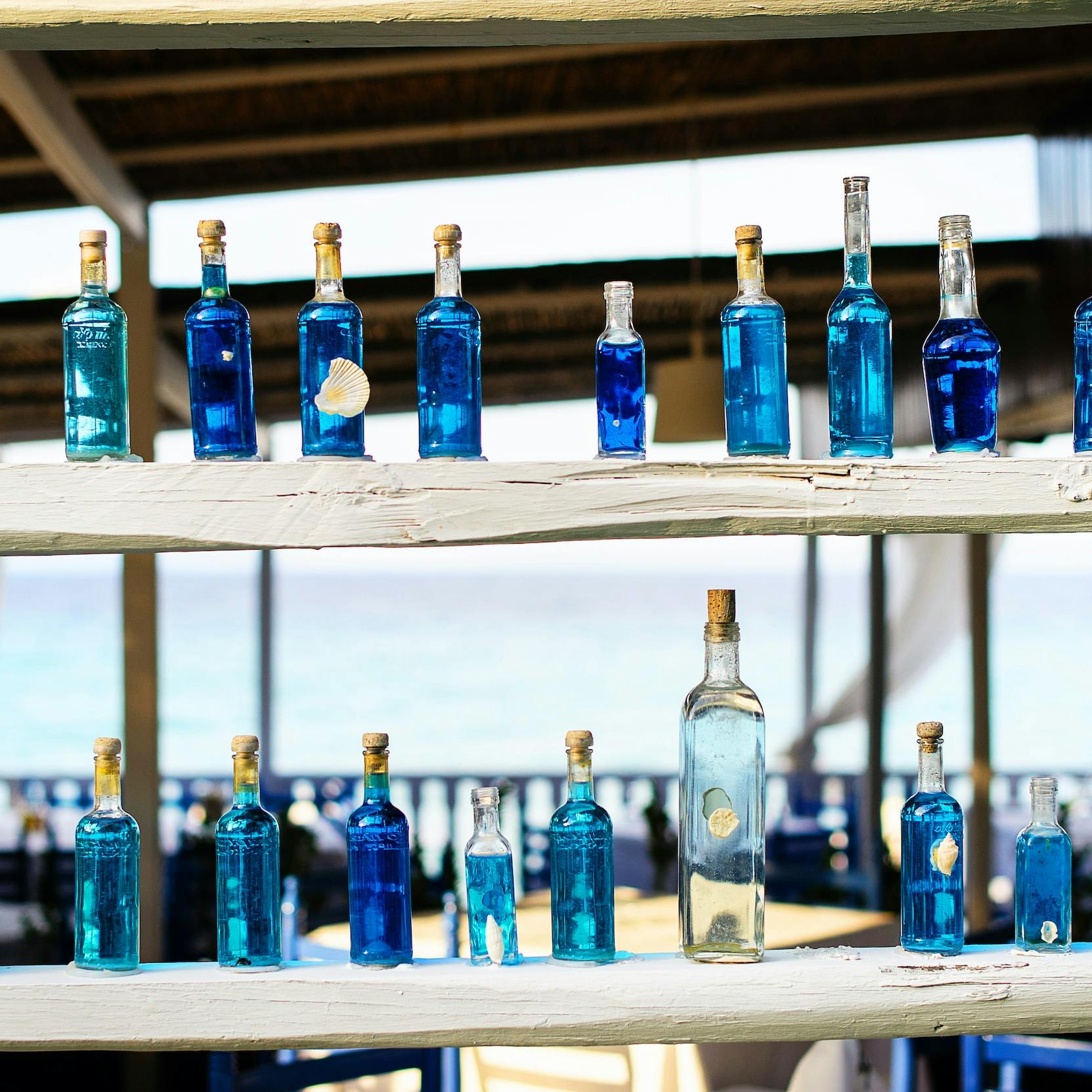 Tante bottiglie di colore azzurro  disposte su due file sopra a delle mensole di legno grezzo, sullo sfondo il mare