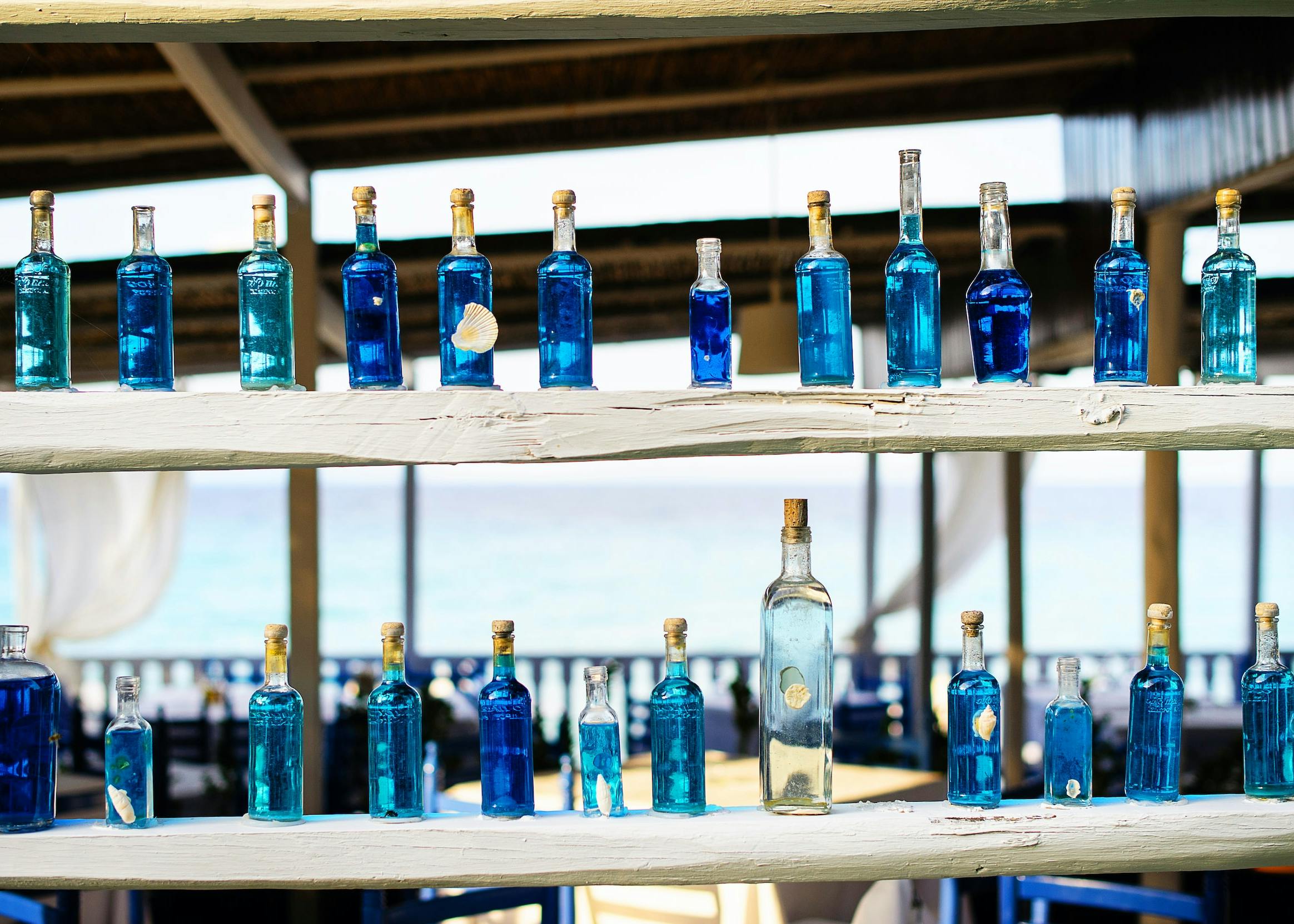 Tante bottiglie di colore azzurro  disposte su due file sopra a delle mensole di legno grezzo, sullo sfondo il mare