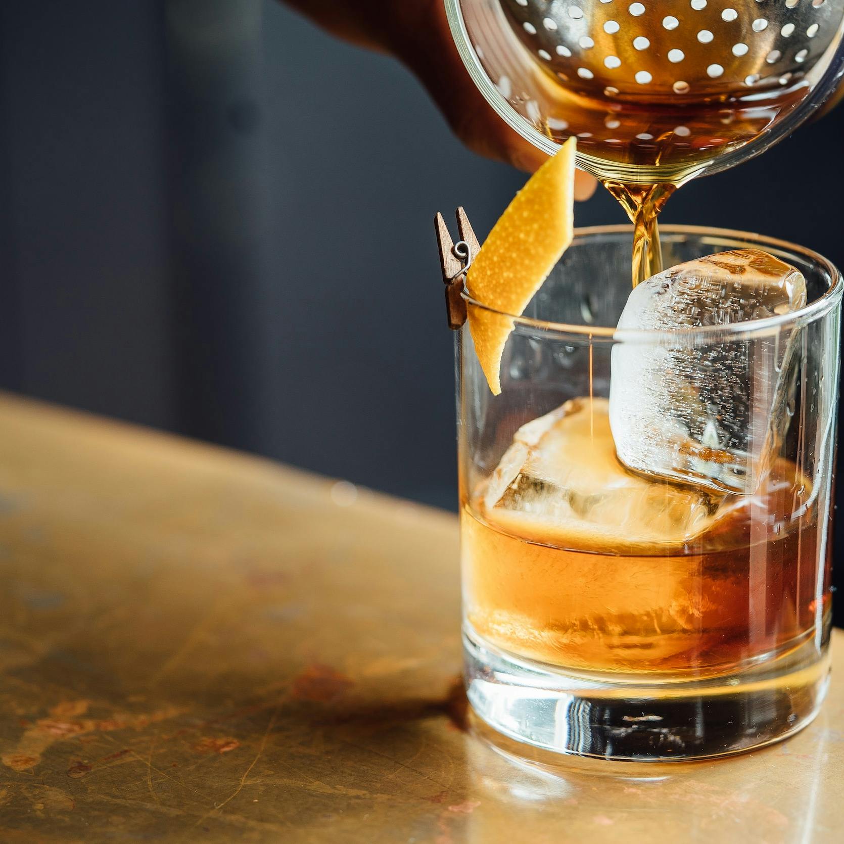 Una mano versa del liquido color ambrato in un bicchiere di vetro, poggiato su un tavolo color oro e pieno a metà, con cubetti di ghiaccio, scorza di arancia fissata al bordo con una moletta di legno.