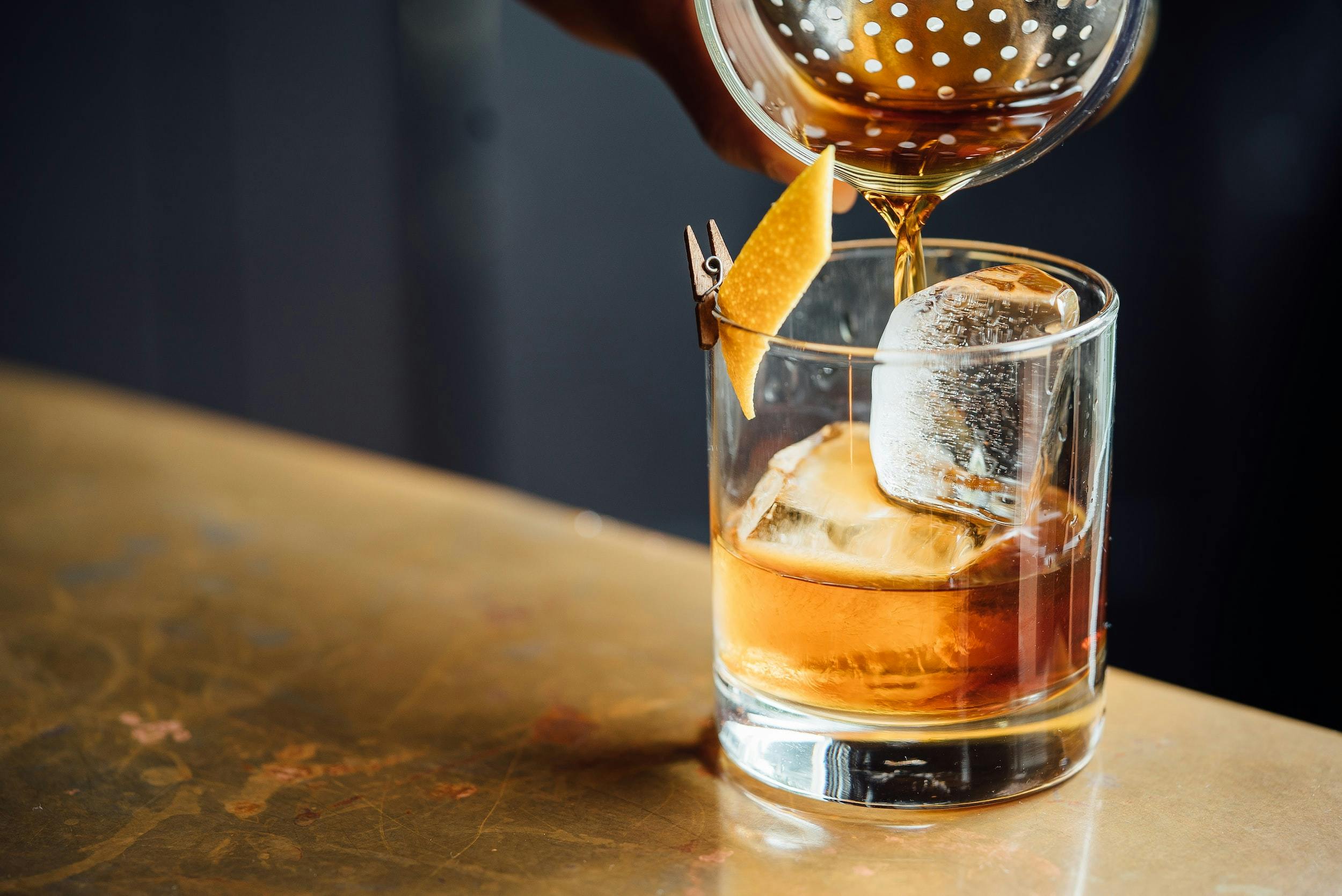 Una mano versa del liquido color ambrato in un bicchiere di vetro, poggiato su un tavolo color oro e pieno a metà, con cubetti di ghiaccio, scorza di arancia fissata al bordo con una moletta di legno.