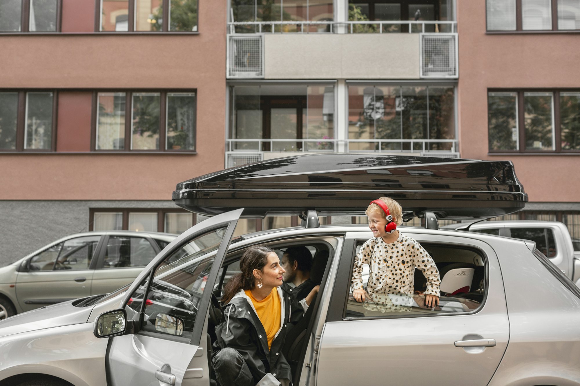 Kvinna i förarsätet med öppen dörr tittar på ett barn med röda hörlurar som ser ut genom ett öppet fönster i baksätet.