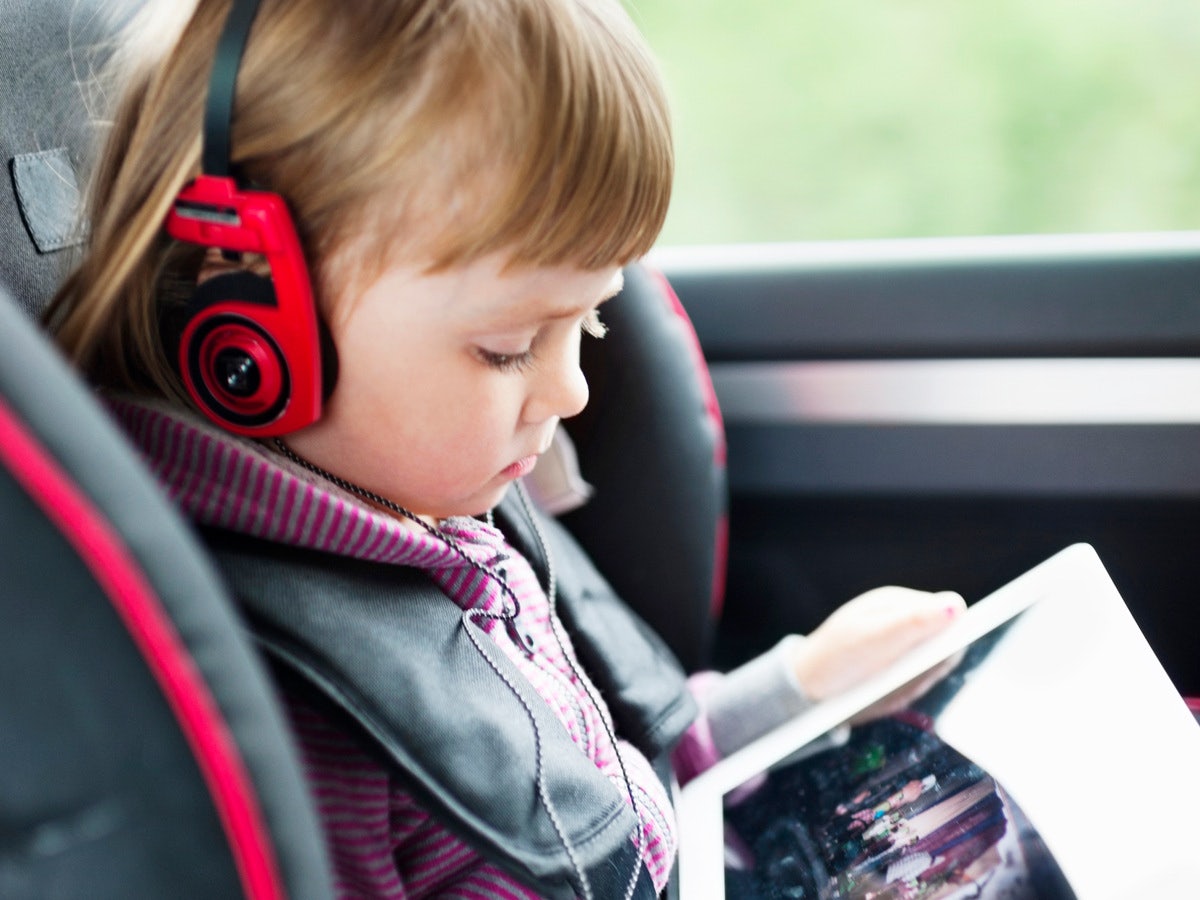 En flicka med röda hörlurar som tittar på en surfplatta i en bilbarnstol.