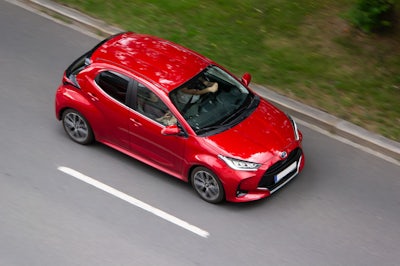 En röd bil kör längs en väg bredvid ett grönskande fält