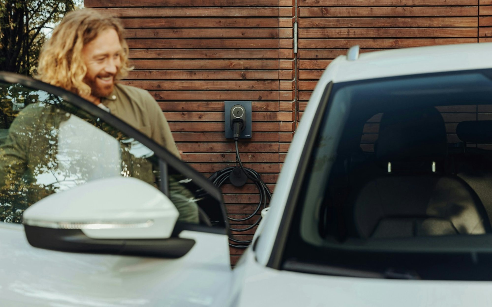 Man står vid en elbil som laddas vid en laddbox monterad på en träfasad och öppnar bildörren.