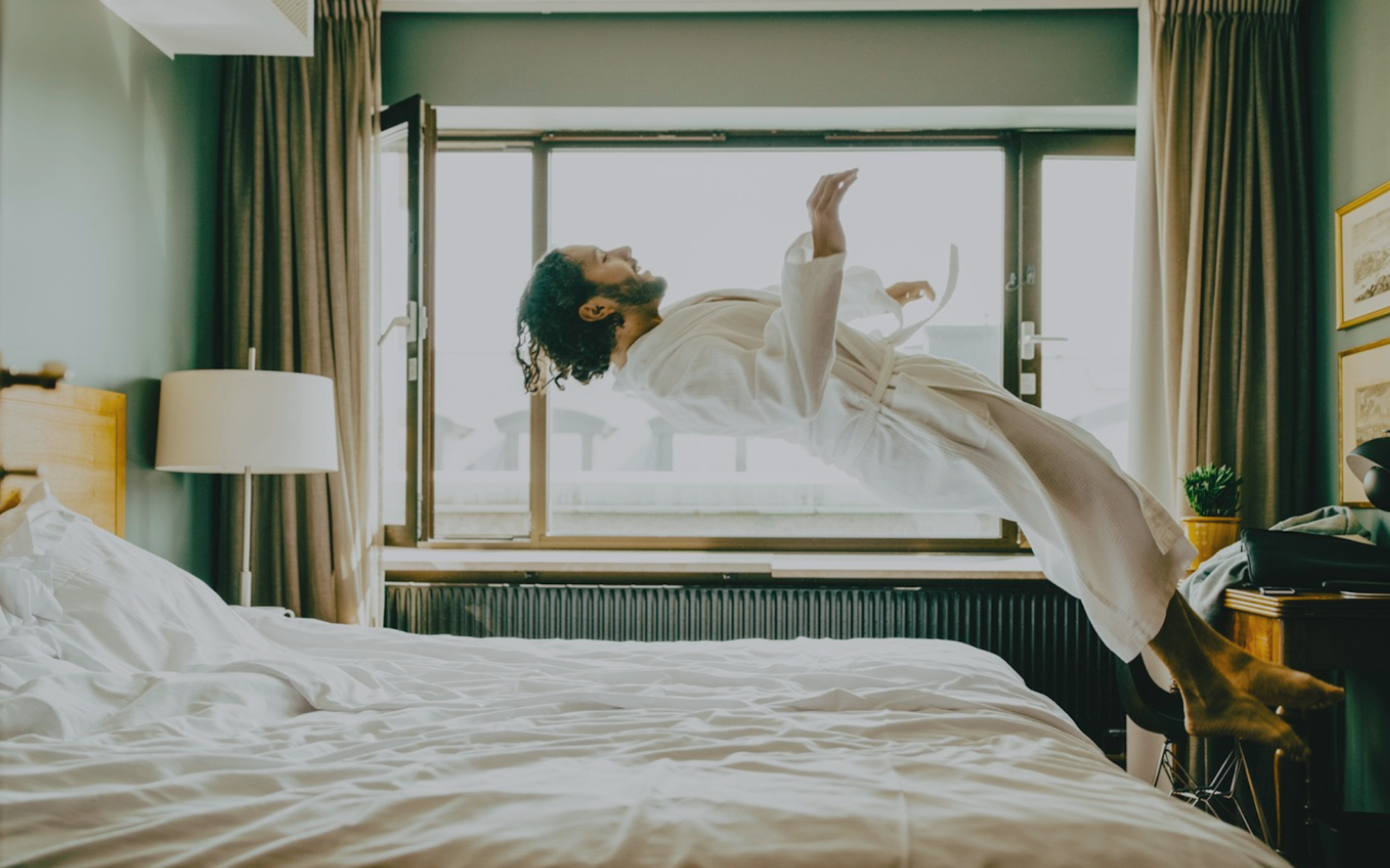 Man i morgonrock svävar ovanför en säng i ett ljust hotellrum.