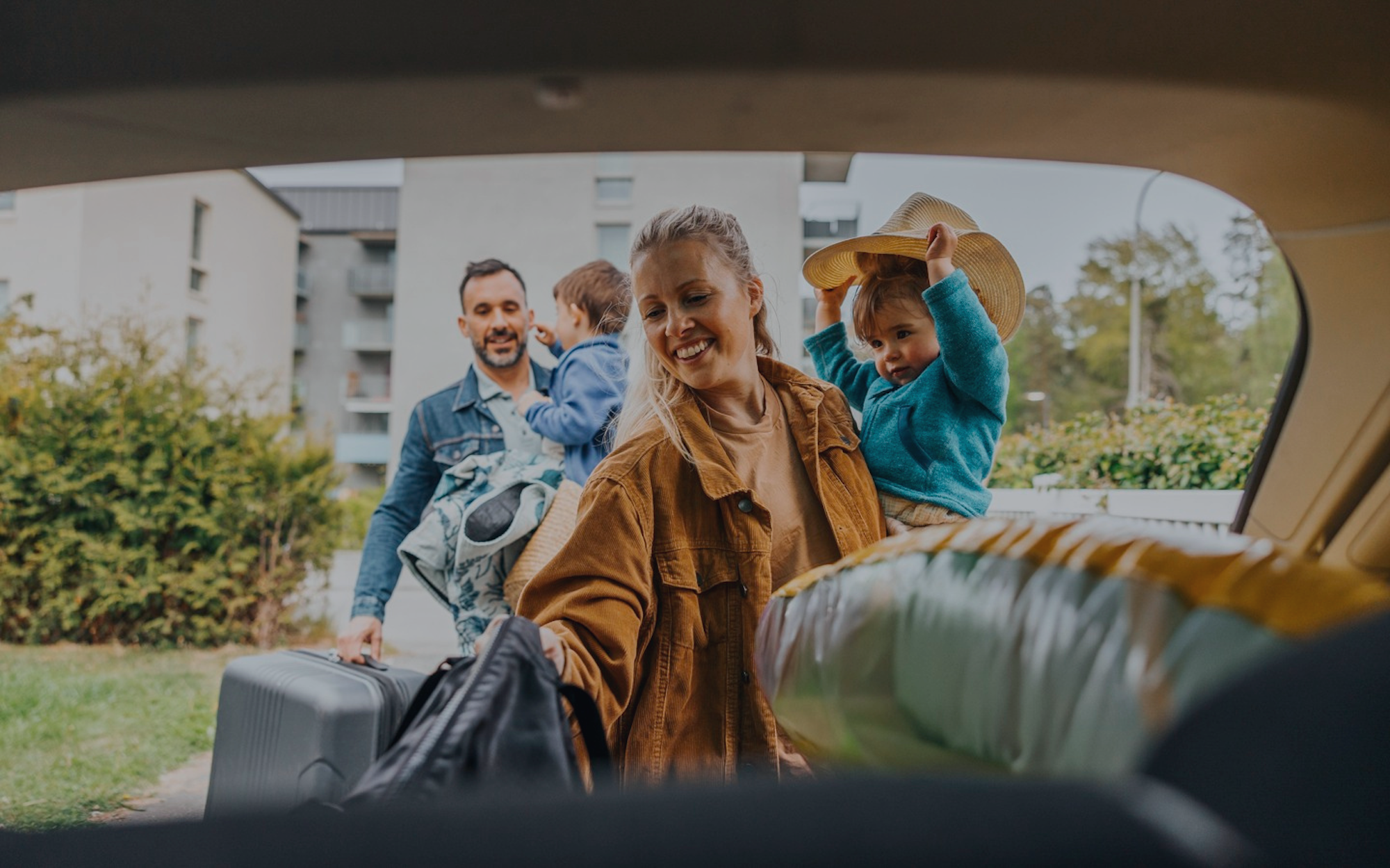 Familj packar in väskor i en bil utanför ett bostadsområde, där en vuxen håller ett barn och en annan bär bagage.