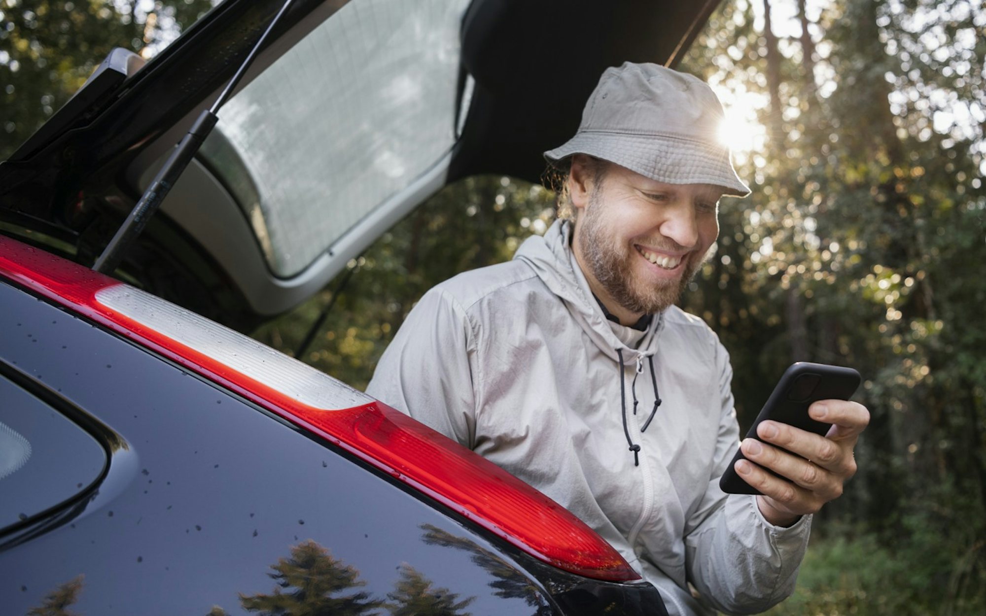 Man sitter i bagaget på en bil i en skog och tittar på mobilen