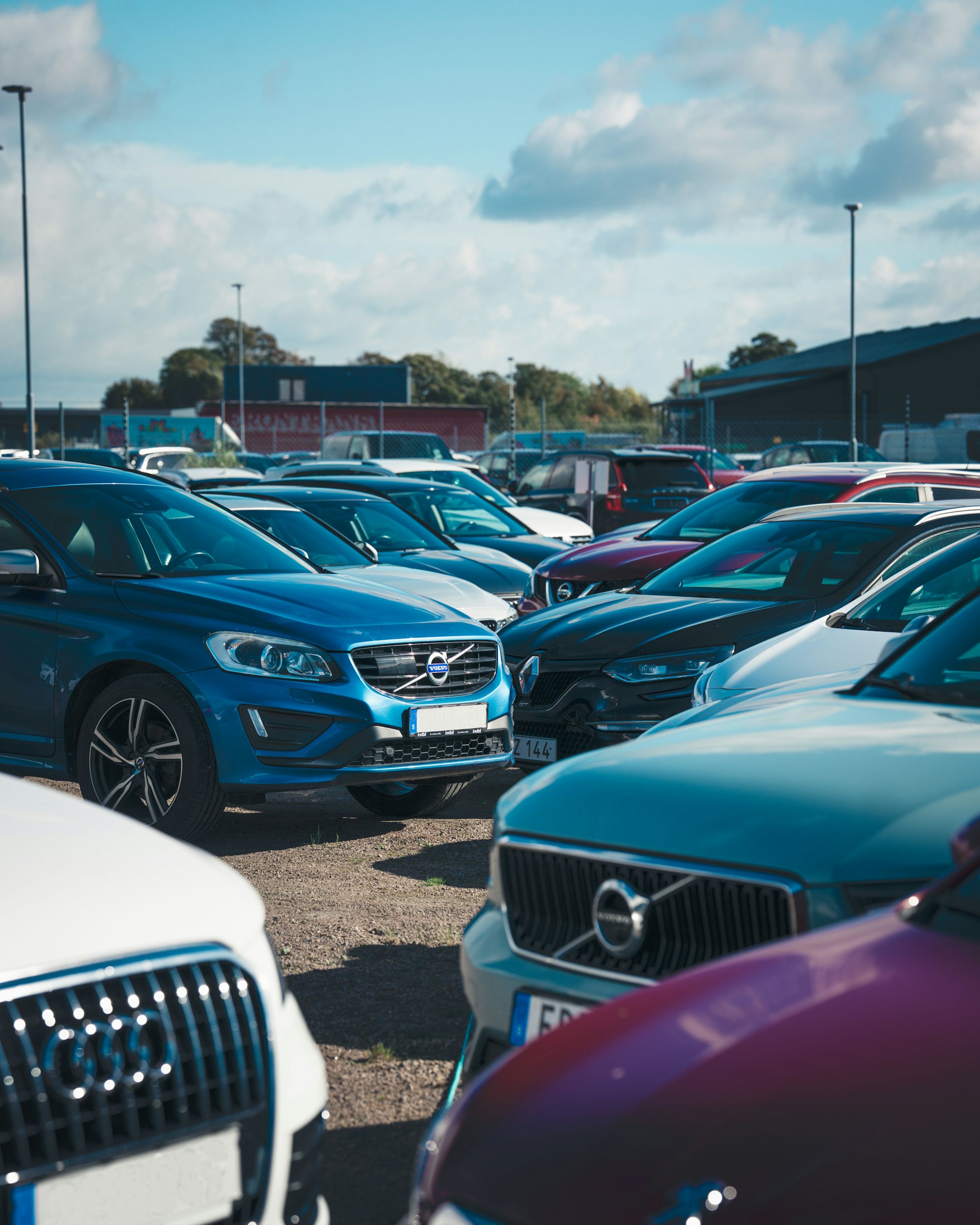 Fullsatt grusparkering med flera Volvobilar, en blå Volvo XC60 i fokus, under blå himmel med moln.