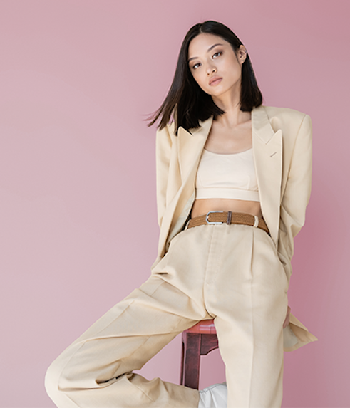 Woman in a cream tailored blazer, matching wide-leg trousers, and a beige crop top with a woven brown belt, seated against a dusty pink background.