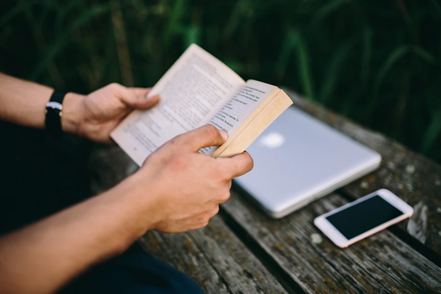 Person holding an open book. Laptop computer and cellphone in background