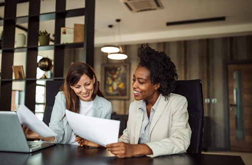 Two ladies at a desk looking at a laptop with paper in their hands