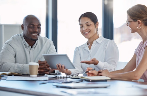 3 people sat around a table looking at a tablet device and smiling