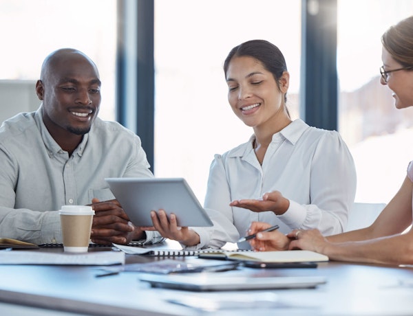 3 people sat around a table looking at a tablet device and smiling