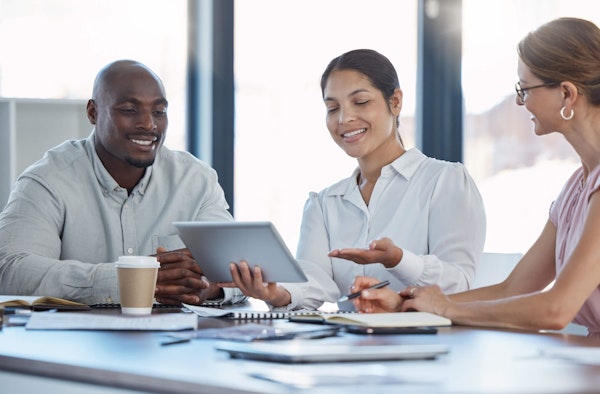 3 people sat around a table looking at a tablet device and smiling
