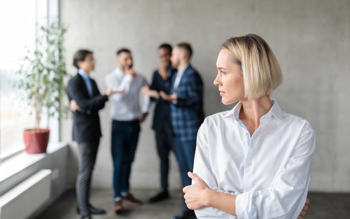 A woman standing alone while a meeting takes place behind her.