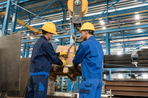 Two workers in hard hats securing a pallet to a crane hook