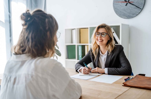 Two women in business attire sat at a desk in conversation