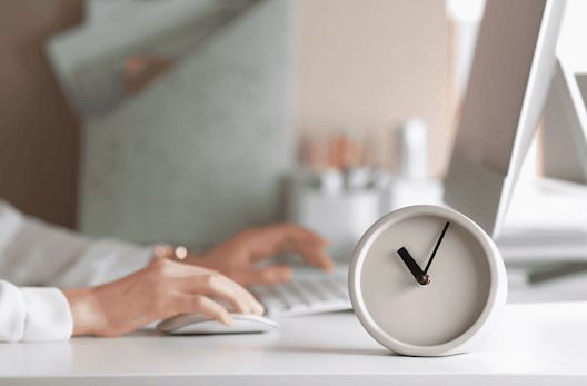 A small desk clock with someone working an a computer in the background