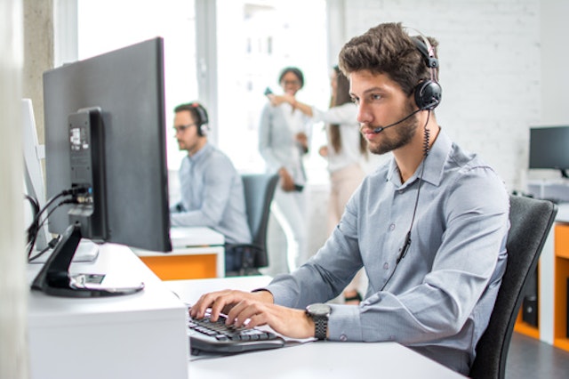 A man wearing a phone headset while sitting at a desk with a computer