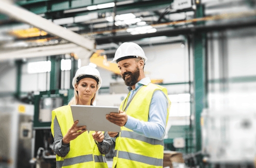 Two construction workers (one man, one woman) wearing high-vis jackets and helmets looking at a tablet.