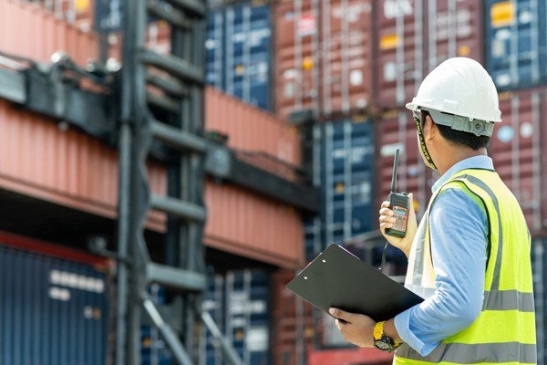 A man in a hard hat and hi-vis talking on a radio surrounded by shipping containers