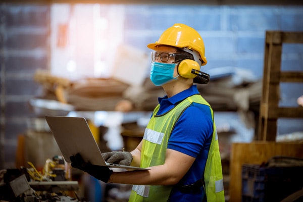 A worker wearing a hard hat and mask, making notes on a laptop