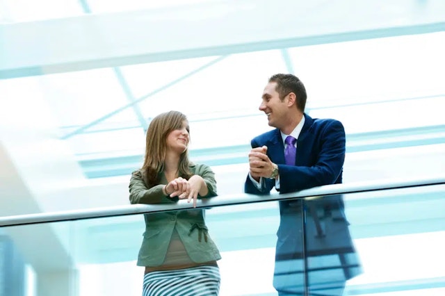 Two colleagues - a man and a woman - are stood on a balcony at work, chatting and smiling.