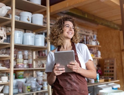 A smiling woman wears an apron and holds a tablet.
