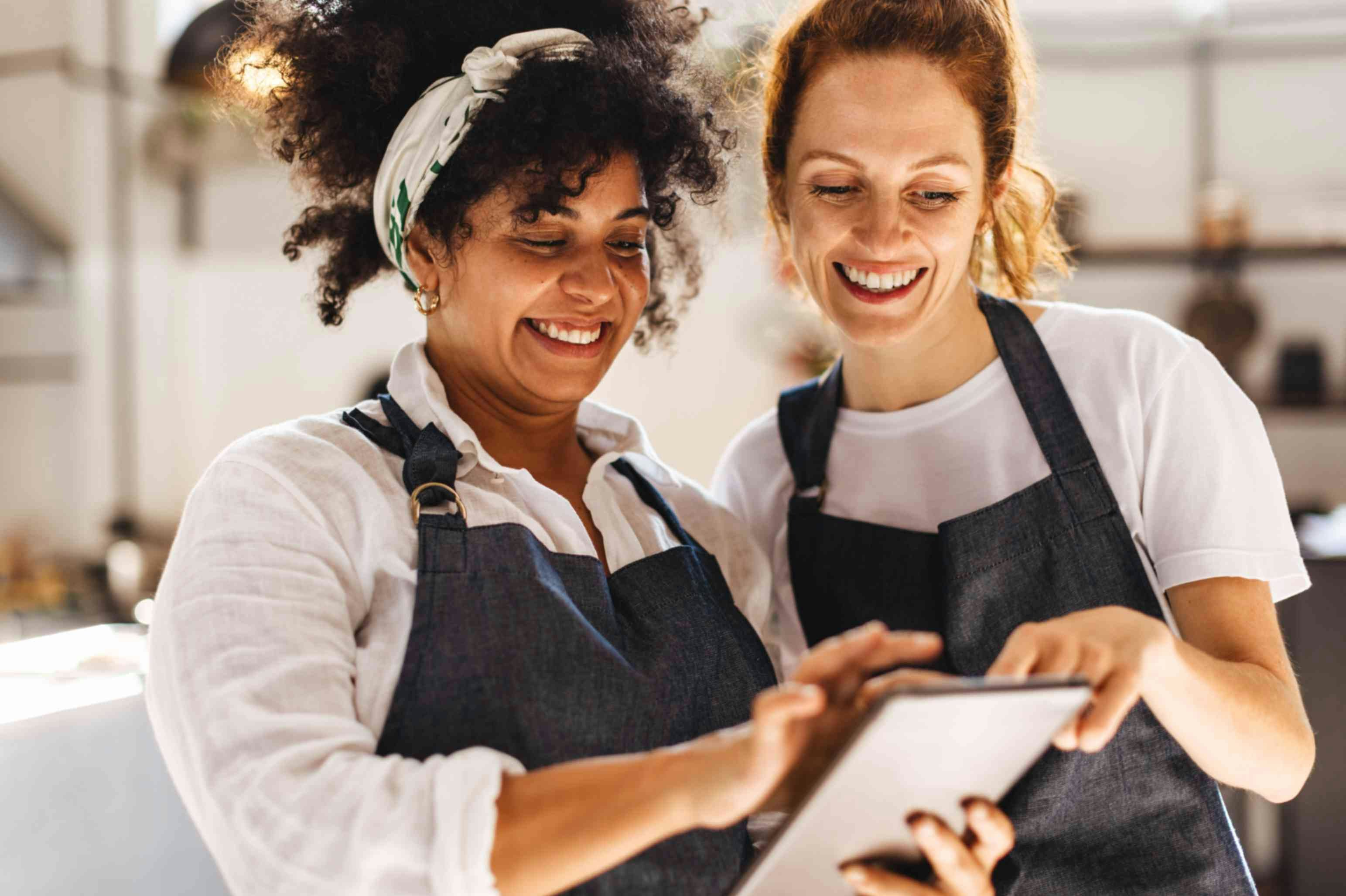 two smiling women wearing aprons looking at a tablet