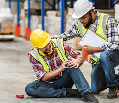 two men on a warehouse floor in hard hats, one is holding his knee
