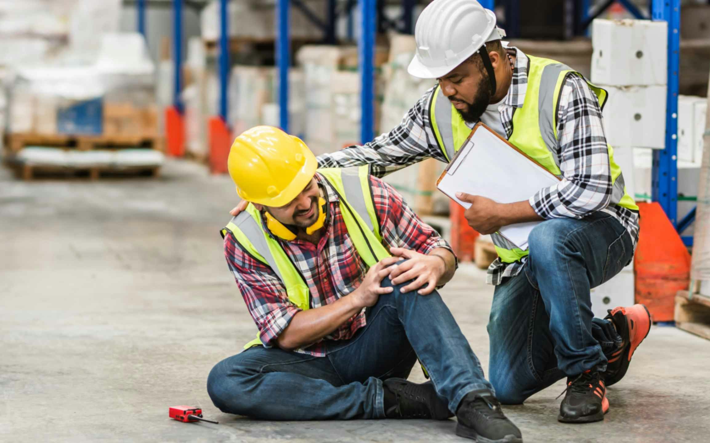 two men on a warehouse floor in hard hats, one is holding his knee