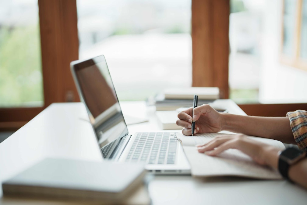a person writing at a desk with a laptop