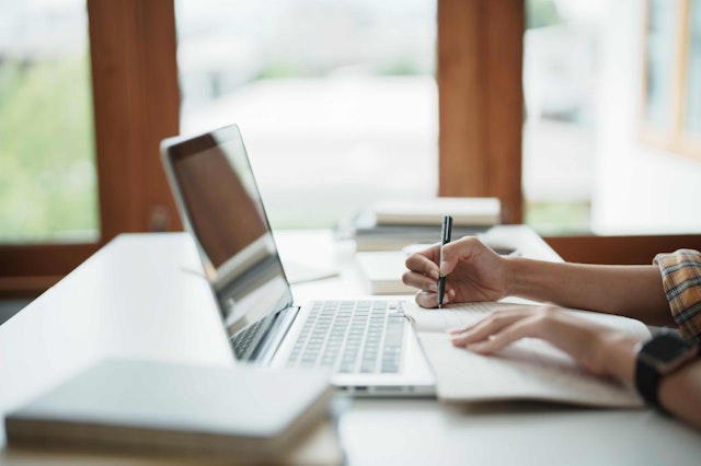 a person writing at a desk with a laptop