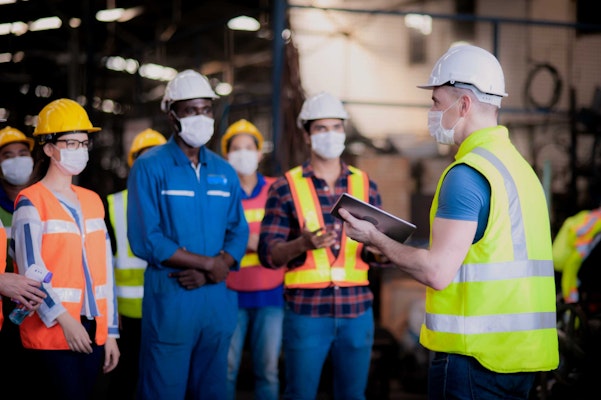 people in PPE following a man with a clipboard and helmet
