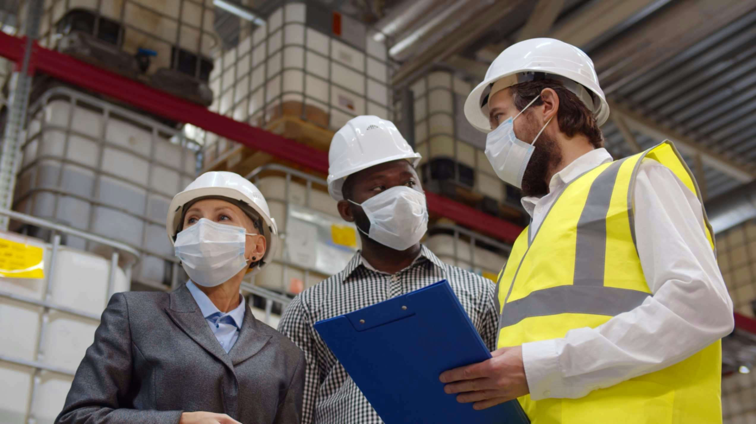 people in a warehouse with hard hats a face masks and a clipboard
