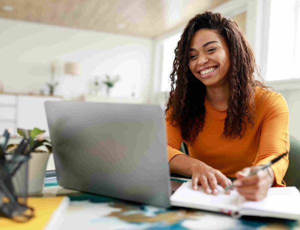 A woman smiles and takes notes while she looks at a screen.