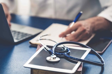 a man writing notes at a desk with a stethoscope and laptop