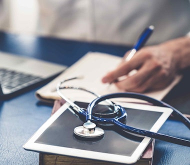 a man writing notes at a desk with a stethoscope and laptop