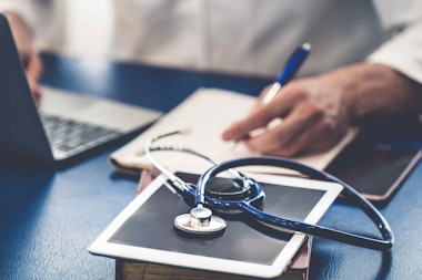a man writing notes at a desk with a stethoscope and laptop