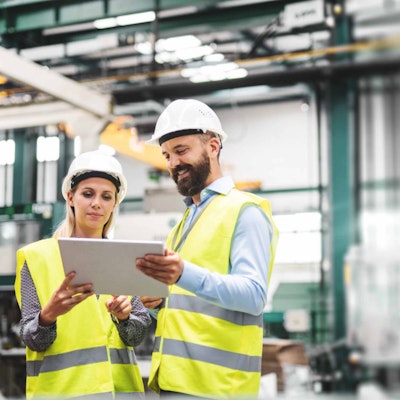 A man and a woman in hard hats and PPE looking at a tablet in a warehouse